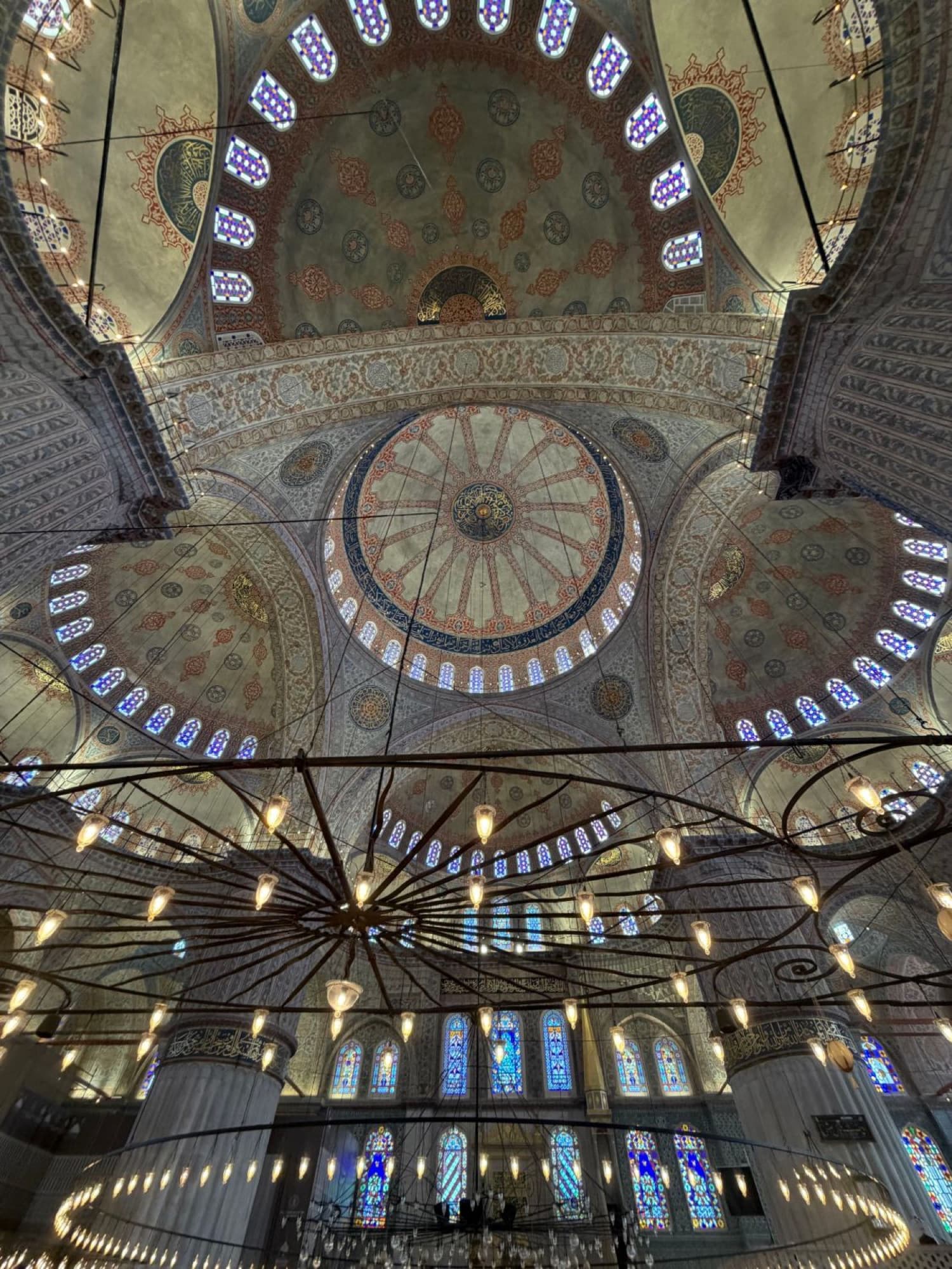 Looking up into the painted domes and stained-glass windows of the Blue Mosque, with a ring of hanging lamps below.