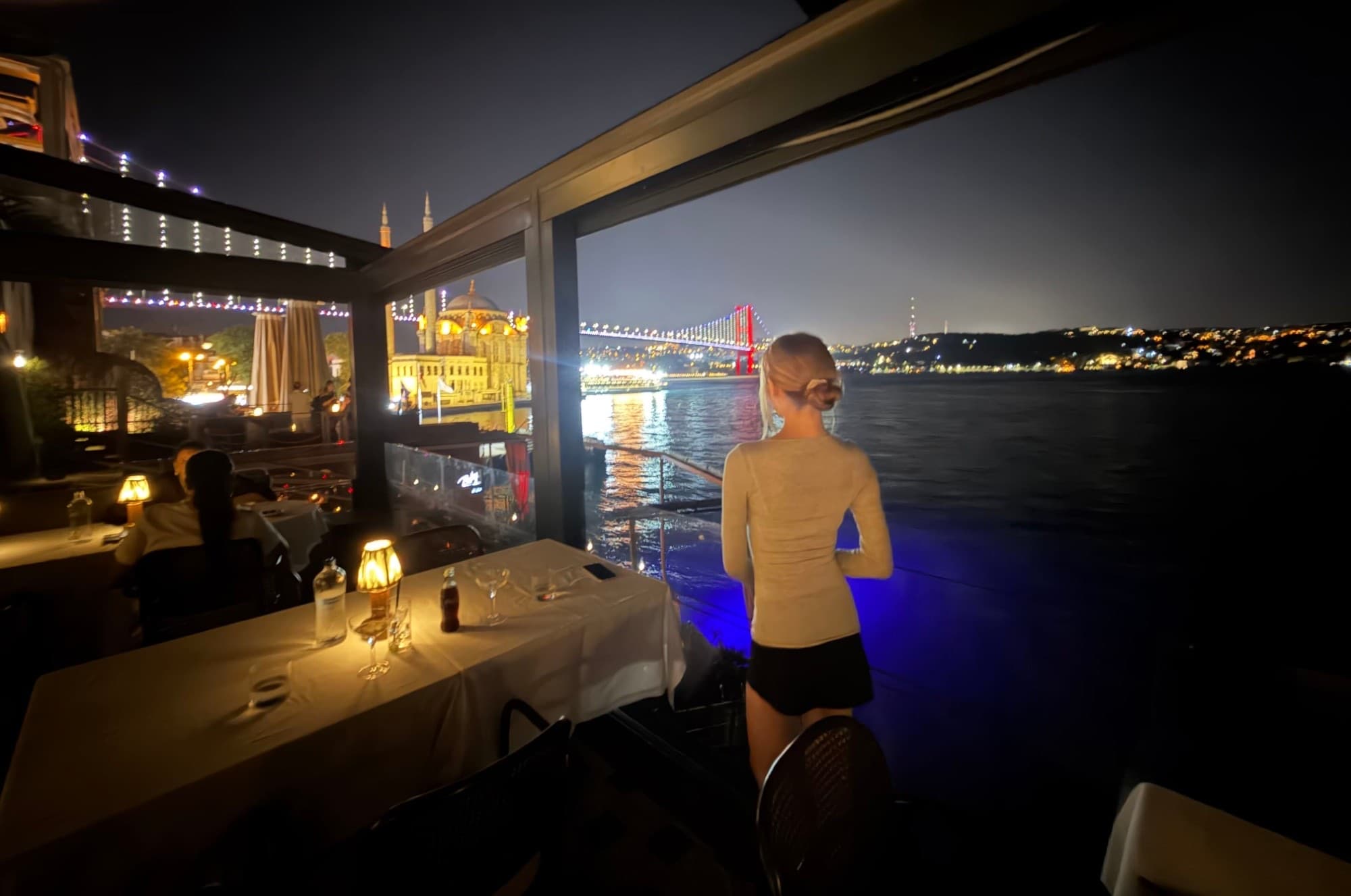 Sofia at a waterfront restaurant at night, looking out over the Bosphorus toward the Ortaköy Mosque and the lit-up Bosphorus Bridge.
