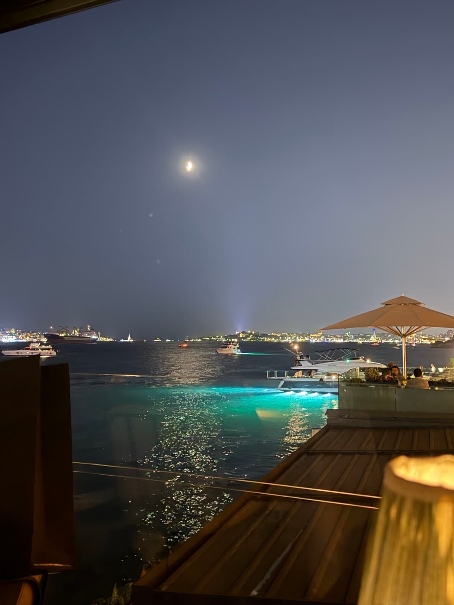 A moon over the Bosphorus at night, with boats on the water and the city lit up along the far shore.
