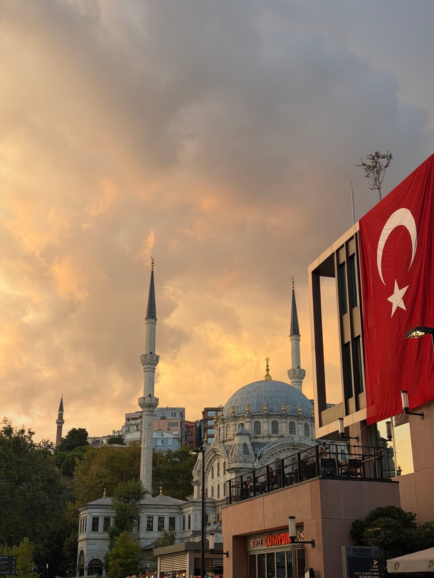 Minarets and the dome of a mosque against an orange evening sky beside Galataport, with a large Turkish flag hanging from a building.